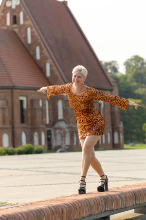 A mature woman balances gracefully on a narrow brick edge outdoors, arms extended like wings. Golden sequin dress and heeled shoes contrast with architectural lines and natural background, creating a vivid lifestyle and fashion portrait.の写真素材