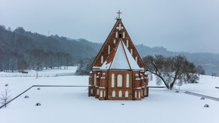 Historic brick church standing alone on the snowy bank of the Nemunas river in Zapyskis, Lithuania during active snowfall. Calm winter landscape, overcast sky, river bend and snow covered trees.の写真素材
