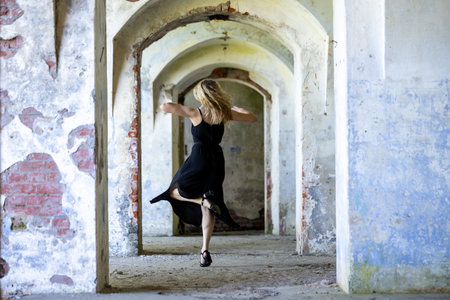 Expressive contemporary dancer in a flowing black dress performing dynamic movements inside an abandoned arched corridor. Concept of freedom, grace, femininity, motion and contrast between body and rough architectureの写真素材