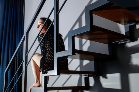 Woman sitting on modern staircase in soft natural light. Minimalist interior, geometric lines and shadows, calm mood, contemporary lifestyle, privacy, solitude, modern home architectureの写真素材
