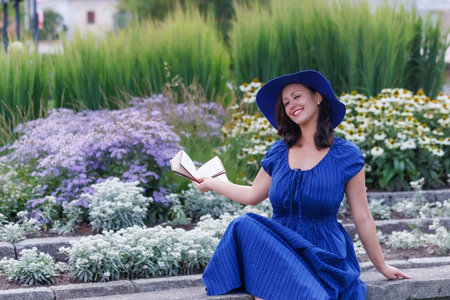 Smiling woman wearing a blue summer dress and wide brim hat, sitting in a flower garden and reading a book. Relaxed lifestyle portrait, leisure time, femininity, nature, copy spaceの写真素材