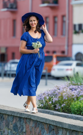 A woman in a blue summer dress walks outdoors on a warm day, holding flowers and a wide-brimmed hat. A cheerful, carefree lifestyle moment in an urban environment, full of light and natural emotionの写真素材