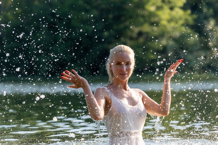 Joyful young woman in a white summer dress enjoying playful water splashes in a natural lake setting on a sunny day. Energetic motion captured in mid-air with sparkling water drops, creating a sense of freedom, freshness, and happiness in nature. Perfect for concepts of summer, vitality, relaxation, and outdoor lifestyle.の写真素材