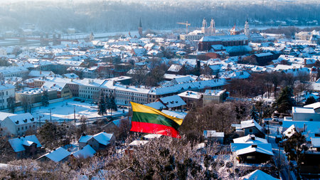 A large Lithuanian tricolor flag flies in the wind above a snow-covered Kaunas cityscape, showing winter rooftops and historic buildings in bright cold sunlight.の写真素材