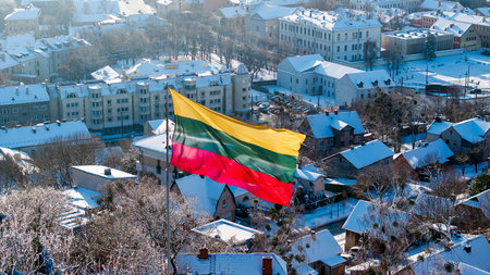 A large Lithuanian tricolor flag flies in the wind above a snow-covered Kaunas cityscape, showing winter rooftops and historic buildings in bright cold sunlight.の写真素材