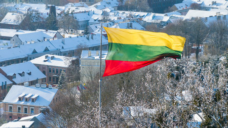 A large Lithuanian tricolor flag flies in the wind above a snow-covered Kaunas cityscape, showing winter rooftops and historic buildings in bright cold sunlight.の写真素材