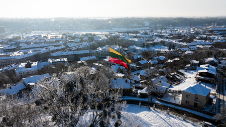 A large Lithuanian tricolor flag flies in the wind above a snow-covered Kaunas cityscape, showing winter rooftops and historic buildings in bright cold sunlight.の写真素材