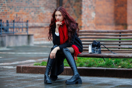 Stylish young woman sitting on a bench in an urban environment on a rainy day. Fashionable outfit with a red scarf, black coat, and knee-high boots creates a cinematic street portrait mood. Urban lifestyle, confidence, and modern city atmosphereの写真素材