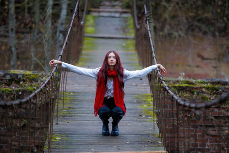 Young woman wearing a red scarf, white shirt, and black pants poses and balances on an old wooden pedestrian bridge surrounded by autumn forest. Conceptual outdoor fashion and lifestyle portrait exploring balance, stillness, and body awareness in a rustic natural environment. Calm, moody atmosphere with natural light and copy space.の写真素材
