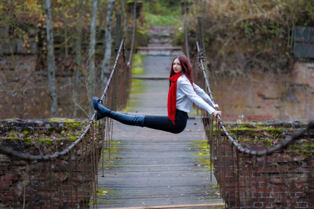 Young woman wearing a red scarf, white shirt, and black pants performs expressive stretching and balancing poses on an old wooden pedestrian bridge surrounded by autumn forest. Conceptual outdoor fashion and lifestyle portrait combining movement, balance, and freedom in a natural rustic environment.の写真素材