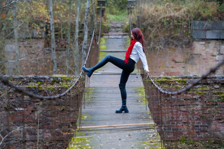 Young woman wearing a red scarf, white shirt, and black pants performs expressive stretching and balancing poses on an old wooden pedestrian bridge surrounded by autumn forest. Conceptual outdoor fashion and lifestyle portrait combining movement, balance, and freedom in a natural rustic environment.の写真素材