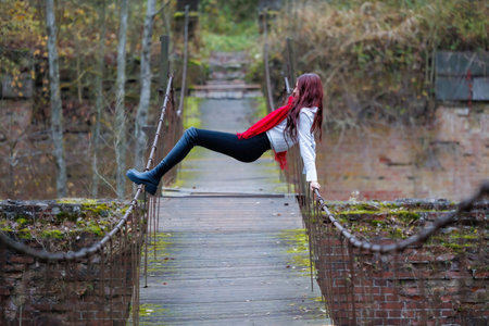 Young woman wearing a red scarf, white shirt, and black pants performs expressive stretching and balancing poses on an old wooden pedestrian bridge surrounded by autumn forest. Conceptual outdoor fashion and lifestyle portrait combining movement, balance, and freedom in a natural rustic environment.の写真素材