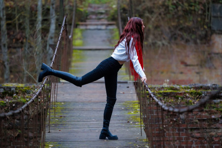 Young woman wearing a red scarf, white shirt, and black pants performs expressive stretching and balancing poses on an old wooden pedestrian bridge surrounded by autumn forest. Conceptual outdoor fashion and lifestyle portrait combining movement, balance, and freedom in a natural rustic environment.の写真素材