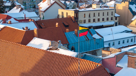 Lithuanian national flag waving above the snowy rooftops of Kedainiai old town during winter. Historic architecture, red tiled roofs and cold seasonal atmosphere in central Lithuania. Patriotic symbol, Baltic country, European cityscapeの写真素材