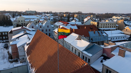 Lithuanian national flag waving above the snowy rooftops of Kedainiai old town during winter. Historic architecture, red tiled roofs and cold seasonal atmosphere in central Lithuania. Patriotic symbol, Baltic country, European cityscapeの写真素材