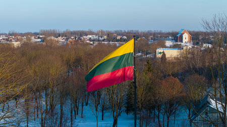 Lithuanian national flag waving above the snowy city of Kedainiai during winter. Urban landscape with residential buildings and trees covered in snow. Patriotic symbol of Lithuania and Baltic region in Northern Europeの写真素材