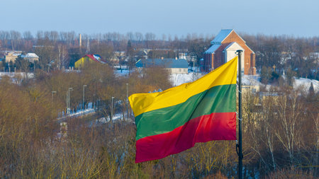 Lithuanian national flag waving above the snowy city of Kedainiai during winter. Urban landscape with residential buildings and trees covered in snow. Patriotic symbol of Lithuania and Baltic region in Northern Europeの写真素材