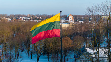 Lithuanian national flag waving above the snowy city of Kedainiai during winter. Urban landscape with residential buildings and trees covered in snow. Patriotic symbol of Lithuania and Baltic region in Northern Europeの写真素材