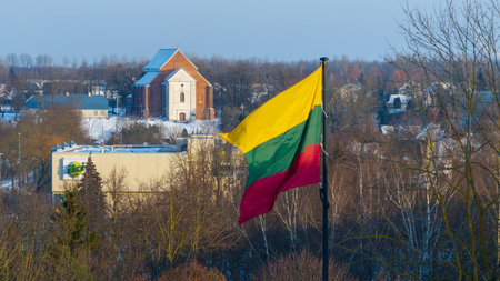 Lithuanian national flag waving above the snowy city of Kedainiai during winter. Urban landscape with residential buildings and trees covered in snow. Patriotic symbol of Lithuania and Baltic region in Northern Europeの写真素材