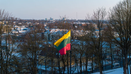 Lithuanian national flag waving above the snowy city of Kedainiai during winter. Urban landscape with residential buildings and trees covered in snow. Patriotic symbol of Lithuania and Baltic region in Northern Europeの写真素材
