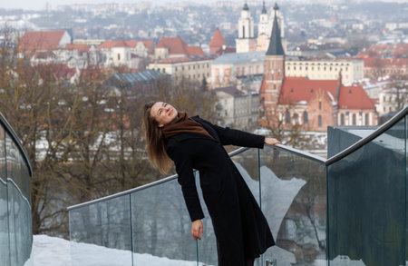 Stylish young woman standing on modern balcony enjoying panoramic winter city view. Concept of independence, confidence, urban lifestyle and personal freedomの写真素材