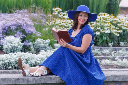 Smiling woman wearing a blue summer dress and wide brim hat, sitting in a flower garden and reading a book. Relaxed lifestyle portrait, leisure time, femininity, nature, copy spaceの写真素材