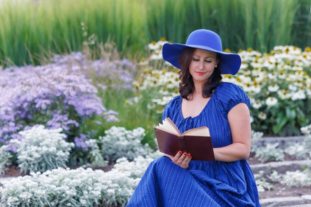 Smiling woman wearing a blue summer dress and wide brim hat, sitting in a flower garden and reading a book. Relaxed lifestyle portrait, leisure time, femininity, nature, copy spaceの写真素材