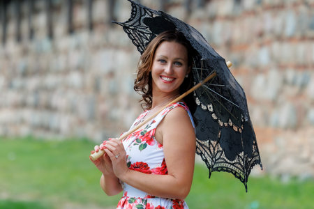 A smiling woman in a floral summer dress stands outdoors with a lace parasol, enjoying a warm and sunny day. A cheerful lifestyle moment with elegance and classic feminine charmの写真素材