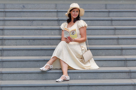 Elegant woman in a light yellow summer dress and straw hat sitting on stone stairs outdoors....