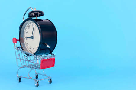Shopping cart and classic alarm clock on blue background. Sale time market shop consumer concept. Selective focus.の写真素材