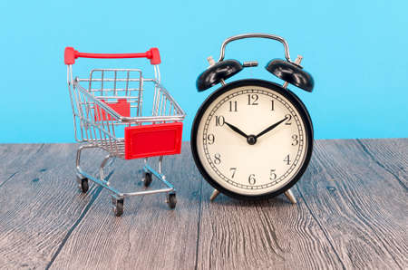 Shopping cart and classic alarm clock on wooden surface. Sale time buy mall market shop consumer concept. Selective focus.の写真素材