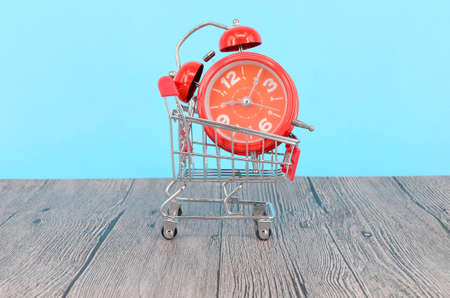 Shopping cart and classic alarm clock on wooden surface. Sale time buy mall market shop consumer...
