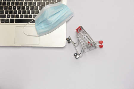 Shopping trolley and medical face mask with laptop on white background. Covid-19 pandemic. Online...