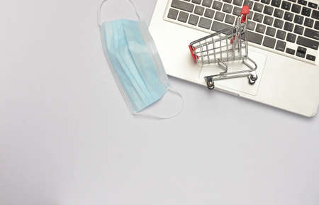 Shopping trolley and medical face mask with laptop on white background. Covid-19 pandemic. Online shopping. Selective focus.の写真素材