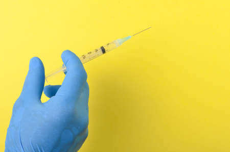 Hand wearing a blue latex glove holding syringe on yellow background. Selective focus.