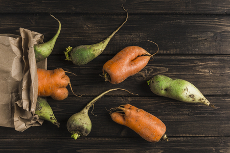 Ugly vegetable green radish and carrot with sprouts on a dark wooden natural background out of a paper bagの写真素材