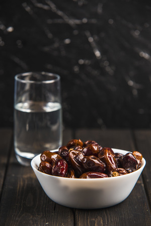 Foods of Ramadan (iftar) with dates, nuts, dried apricots, dried plums in a white bowl with a glass of water on a dark wooden background, side view.の写真素材