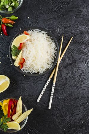 Rice noodles funchosa with vegetables in a black bowl with chopsticks on a dark background, top view, flatlay. Asian food. Copy space.の写真素材