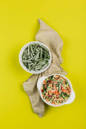 Frozen vegetables: string beans and a mix of vegetables in white plates with a napkin on a bright yellow background. Top view with a copy space. Vertical orientation.の写真素材