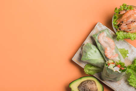 Spring rice paper roll with shrimp, avocado, paprika, lettuce and cottage cheese on a wooden white Board on an orange background. Vegan food. Top view with a copy space. Horizontal orientation.の写真素材