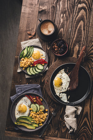 Two plates and a frying pan with cooked eggs with avocado, paprika, cucumber and canned corn on a dark wooden background. Healthy breakfast. Top view with copyspace. Flat lay.の写真素材