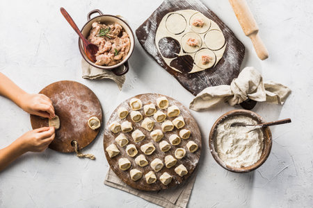 The process of making dumplings - children's hands make dumplings. A lot of ready to cook pork dumplings on a large wooden board with flour on a light background. Top view with copyspace. Horizontal orientation. Flat lay.の写真素材