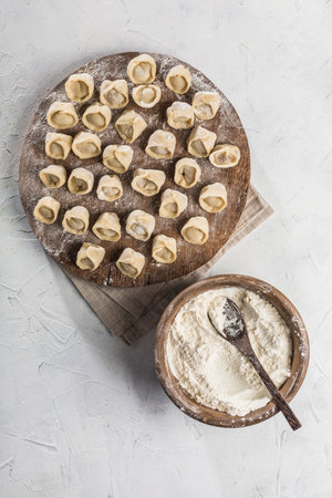 The process of cooking dumplings of Russian and Ukrainian national food. Lots of ready-to-cook pork dumplings on a large wooden board with flour in a bowl on a light background. Top view with copyspace. Vertical orientation. Flat lay.の写真素材