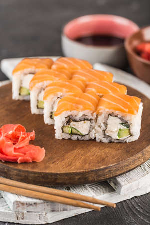Classic Philadelphia sushi rolls on a wooden board with ginger, soy sauce and chopsticks on a dark background. Close-up, selective focus. Vertical orientation.の写真素材