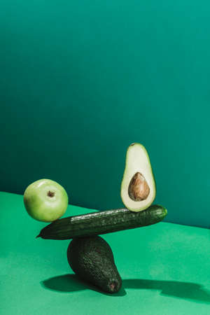 the concept of a balance of fresh vegetables and fruits, cucumber, avocado and apple on a green background. vertical orientation with a copy space for the text.の写真素材