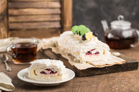 Meringue roll with berry filling with a cut piece on a plate, with a teapot and a mug of tea on a dark background. Side view with a copy space for the text. Dessert for the morning tea party.の写真素材