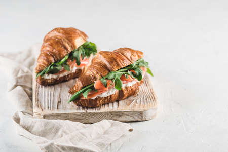 Two croissant sandwich with salmon, ricotta cheese and arugula on a wooden board with a napkin on a light background. Side view with a copy space for the text. Horizontal orientation.の写真素材