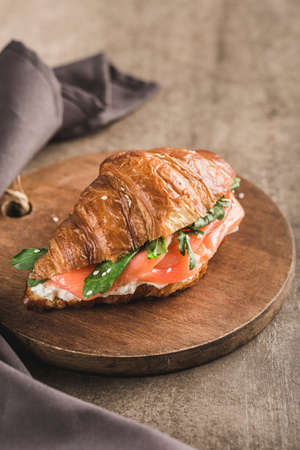 One croissant sandwich with salmon, ricotta cheese and arugula on a wooden board with a napkin on a dark background. Side view with a copy space for the text. Vertical orientation.の写真素材