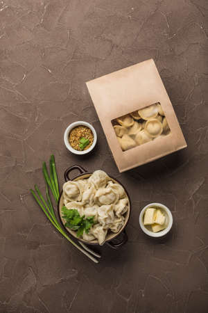 Dumplings in eco-friendly paper packaging for delivery and a bowl with dumplings, onions and sauces on a dark background. Top view with a copy space for the text. Vertical orientation.の写真素材