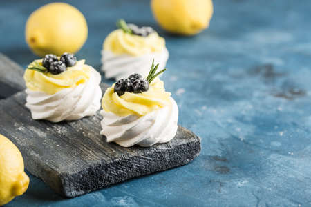 Pavlova dessert with lemon cream and blueberries on a wooden board with lemons on a blue background. A delicious cake for your birthday and holiday. Side view with a copy space for the text. Horizontal orientation.の写真素材
